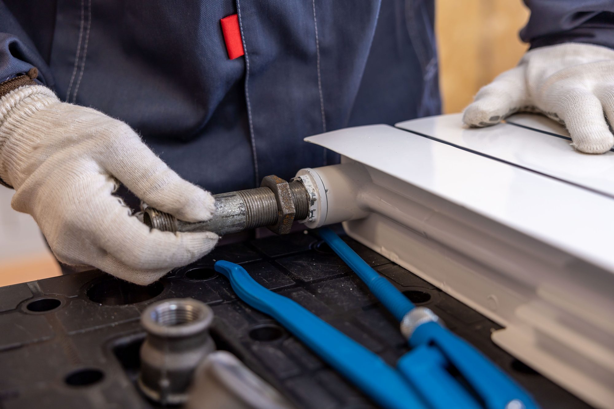 Plumber in gloves fitting a threaded pipe connector to a white radiator, pipe wrenches beside